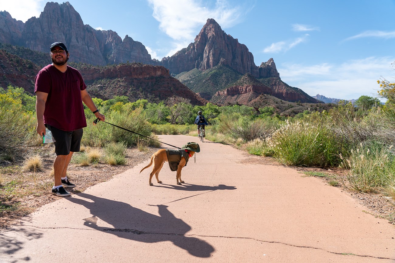 The Pa'rus Trail is pet friendly A man in a red shirt and shorts holds the leash of a dog on a paved path, a bike rider and views of Zion are in the background.