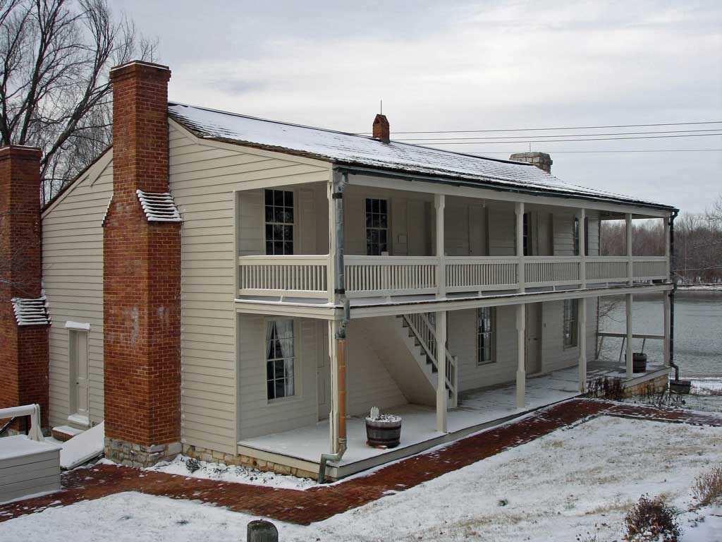 Two story beige building with a front porch on the first and second floor with two red brick chimneys on the side of the building.