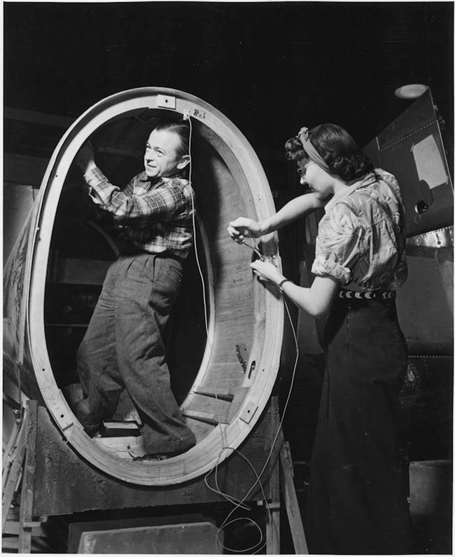 Black and white photo of a little person and a woman installing wires on a plane.  The little person installs wires from within a large oval part, while the woman works from the outside.