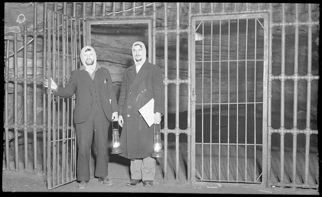 Two men wearing coats and holding lanterns stand near a large iron gate leading into a cave.