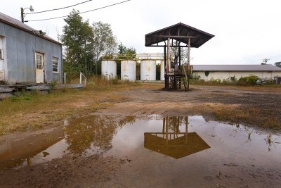 Large brown puddle with abandoned gas pumps, 4 white tanks in the background, weathered blue building on left.