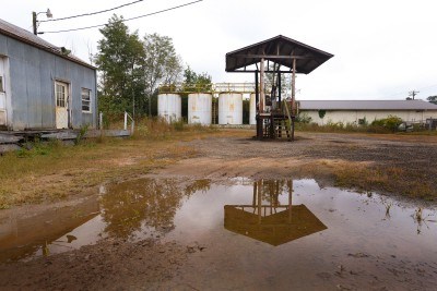 Duncan Oil Company Large brown puddle with abandoned gas pumps, 4 white tanks in the background, weathered blue building on left.