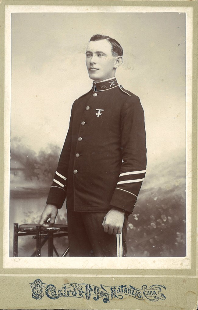 Dunnigan1900 A black and white photo printed on a card. The photo shows a clean-shaven man wearing a military uniform posing in a photo studio.