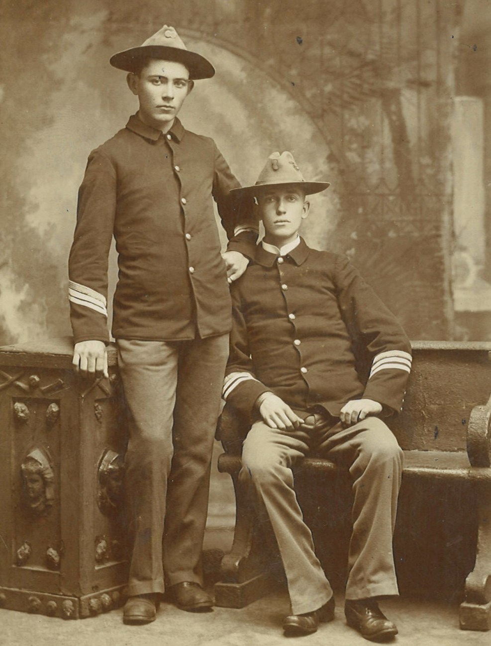 Dunniganandsoldier1898 A black and white image of two men in a portrait studio wearing military uniforms. Christopher Ambrose Dunnigan is a young man with dark hair.