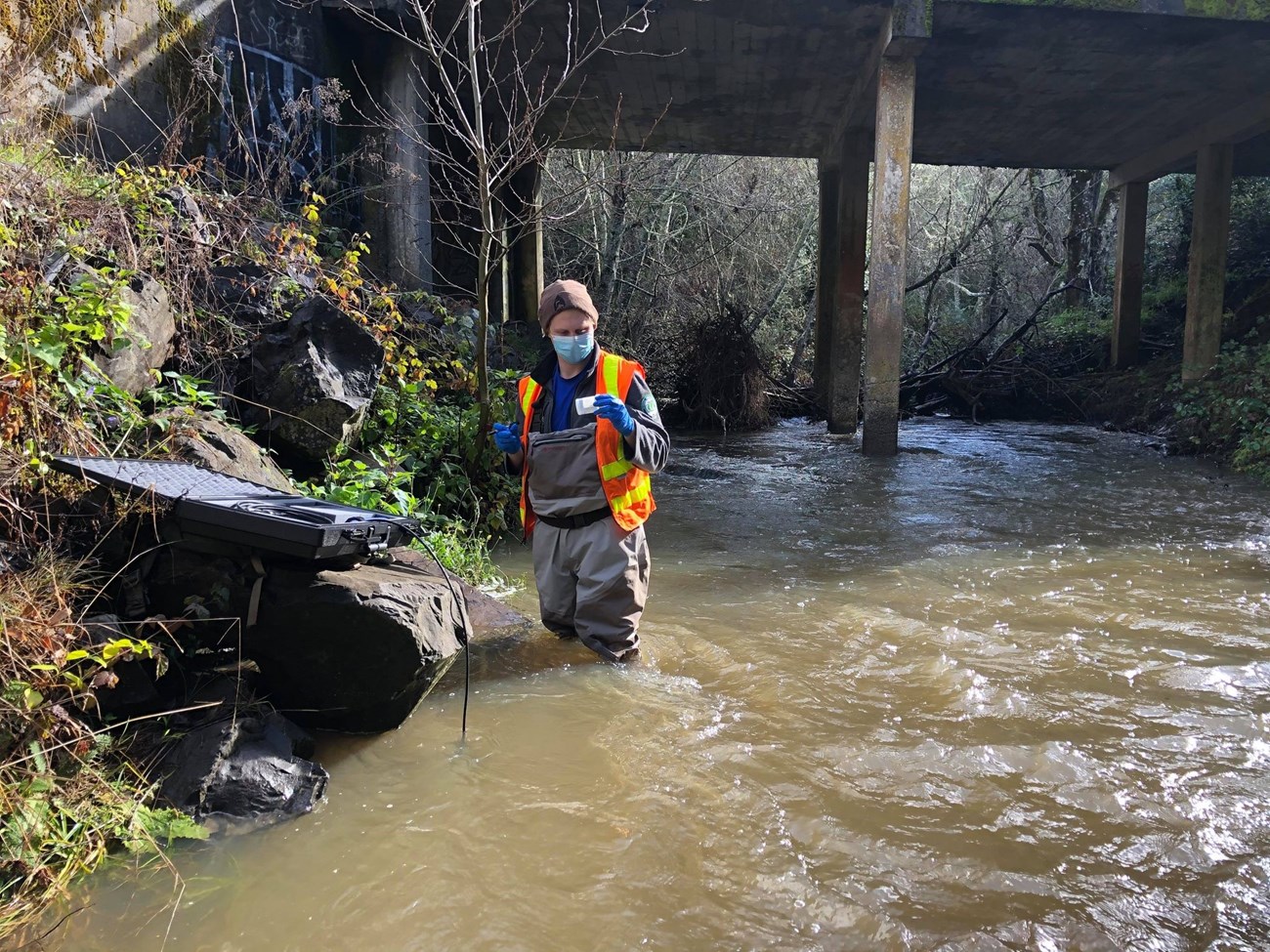 Dustin Geisen Person in waders and a safety vest standing knee-deep in a briskly flowing creek, collecting a water sample.