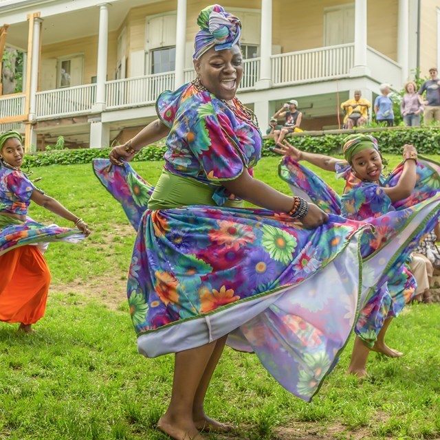A group of dancers in bright skirts dance in front of a yellow house.