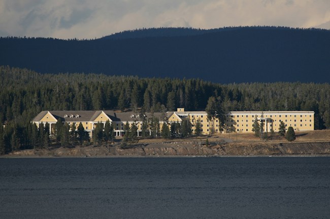 Photograph taken from the water of a large yellow hotel along a lake coastline. The blue lake is in the foreground and a forest is in the background.