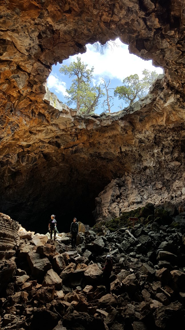 Two cavers stand in the light coming from a large, circular opening in the rock ceiling far above them. The cave floor is covered with rubble of different sizes.