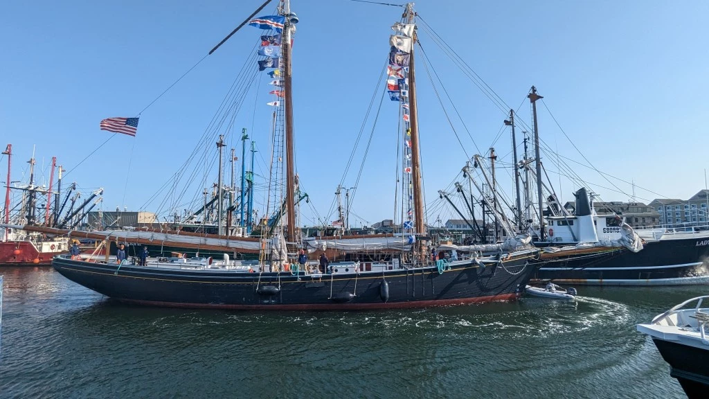 Ernestina-Morrissey returning to New Bedford State Pier in 2023 Schooner with two towering masts and sails lowered in a port surrounded by other vessels.