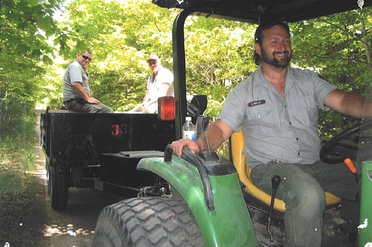 A smiling man in uniform drives a tractor that is pulling a cart with two people on board.