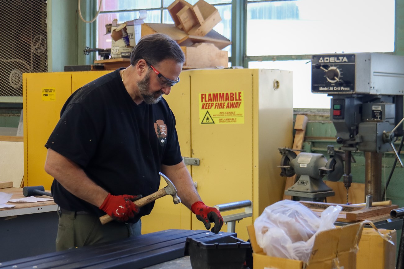 A man working in a shop holds a hammer while looking down at a series of boards.