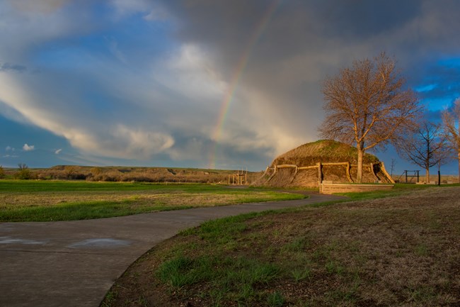 A winding gray path leads to a brown earth lodge. The sky is bright blue and there is a rainbow dropping down on the lodge.