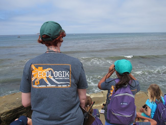 Back of woman wearing a grey t-shirt with an orange octopus and lettering that reads EcoLogik on the back sits next to two girls. All three gaze at the ocean next to Cabrillo National Monument, one of the girls looks through a pair of binoculars.