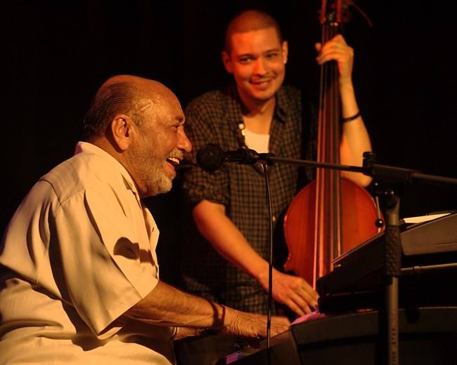 Side view of Eddie Palmieri smiling while he plays the keyboard and sings during a live performance. A young man plays the bass in the background