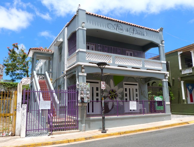 Two-story apartment house painted light blue with white trim. The facade features two projecting balconies with front openings that are framed by an upper horizontal beam.