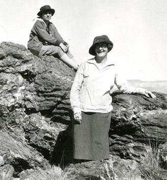 Two women wearing hats face us. One stands leaning on a rock and they second women sits on the rock just behind to the left of the front woman.