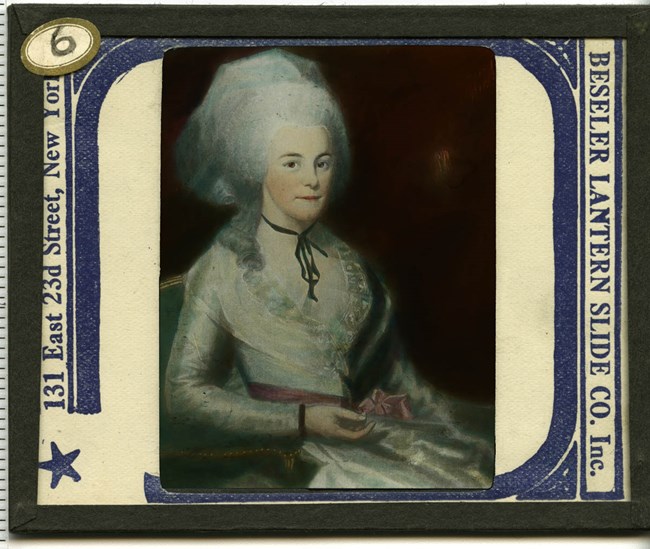 A painted portrait of Elizabeth Schuyler Hamilton, who sits in a silver dress with a large white hairdo. She looks to be in her 20's and smiles slightly.