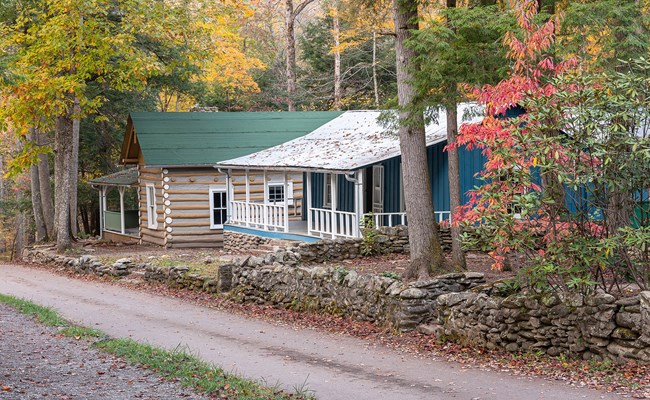 Elkmont cabins_Susan Milinkovich Two historic cabins along a wooded, single-lane road lined with a low rock wall. The first cabin is blue with a white metal roof and white front porch railing. The second has a green roof with log siding.