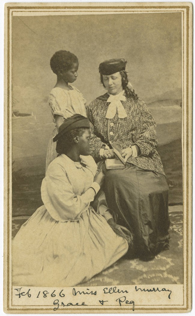Ellen Murray sits in a chair with a book next to two students. “February 1866 Miss Ellen Murray Grace & Peg.”