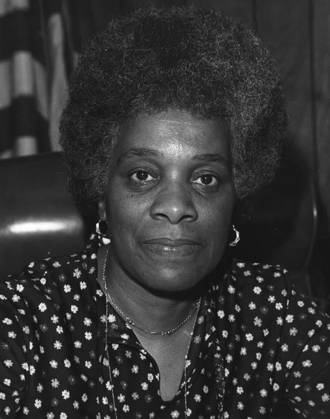 Georgia Ellard Black and white portrait of Georgia Ellard wearing a dark blouse with a flower pattern. US flag behind her.