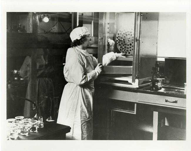 Woman wearing laboratory gown, gloves, and scrub cap works in a laboratory. She stands next to a lab table and reaches into a metal cabinet with a glass front toward a large metal disc with four rows of holes.
