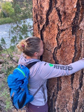 A woman wearing a backpack and a shirt with ‘I Heart RIVERS’ on the sleeve, pressing her nose to the inside of a Ponderosa pine as she hugs the tree.