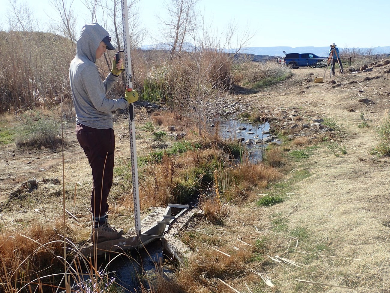 Two scientists at a desert spring - one holds a leveling rod on a flume and another looks through a digital level to check gaging station level.