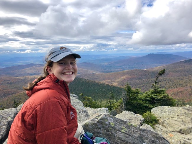 Woman sitting on high rocky point, smiling at camera, with tree-covered hills and mountains behind her.