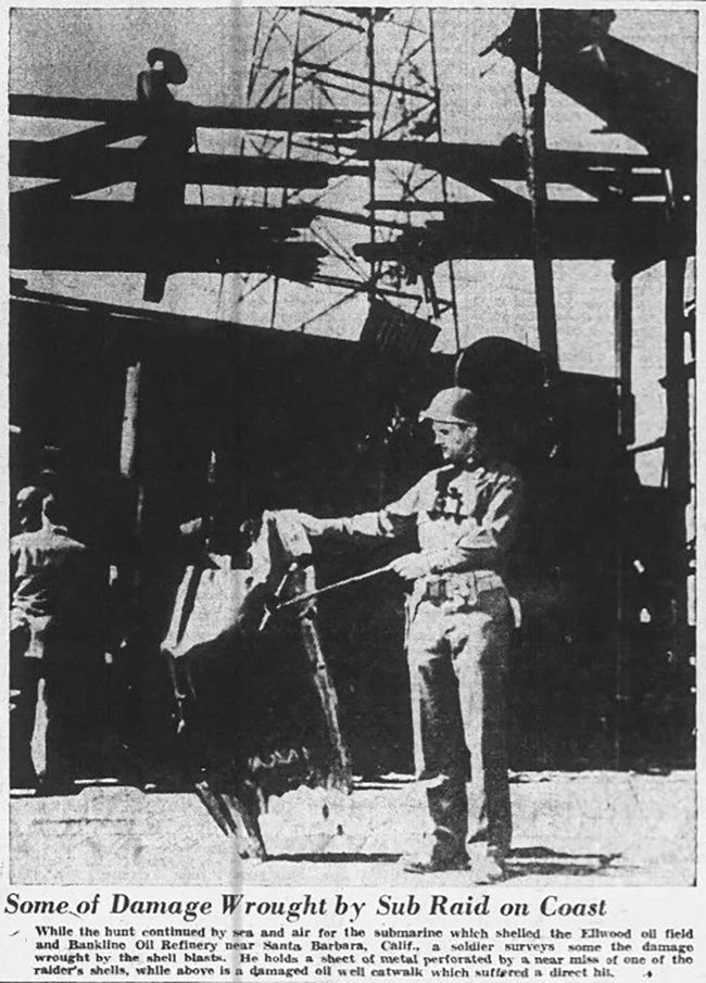 Black and white newspaper photo. In the foreground, a soldier holds a piece of metal wreckage. Above him, another soldier inspects damage to a walkway that has had a hole blown through it.
