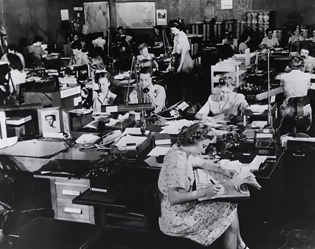 Black and white phot of a room full of desks with mostly white women seated at them, working. There is at least one man present.