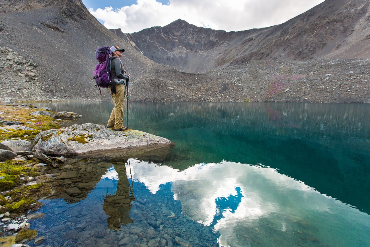 Hiker enjoying an alpine lake Hiker with purple backpack stands on the edge of a small blue green lake with jagged grey peaks in the background