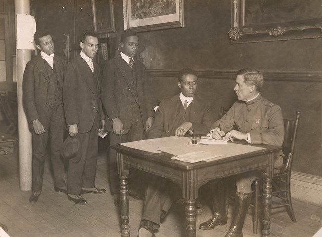 Three Black men wearing suits stand in line behind a fourth Black man. This man is also wearing a suit and sitting next to a white man in uniform behind a square wooden table with loose paper on it. The two seated men make eye contact.