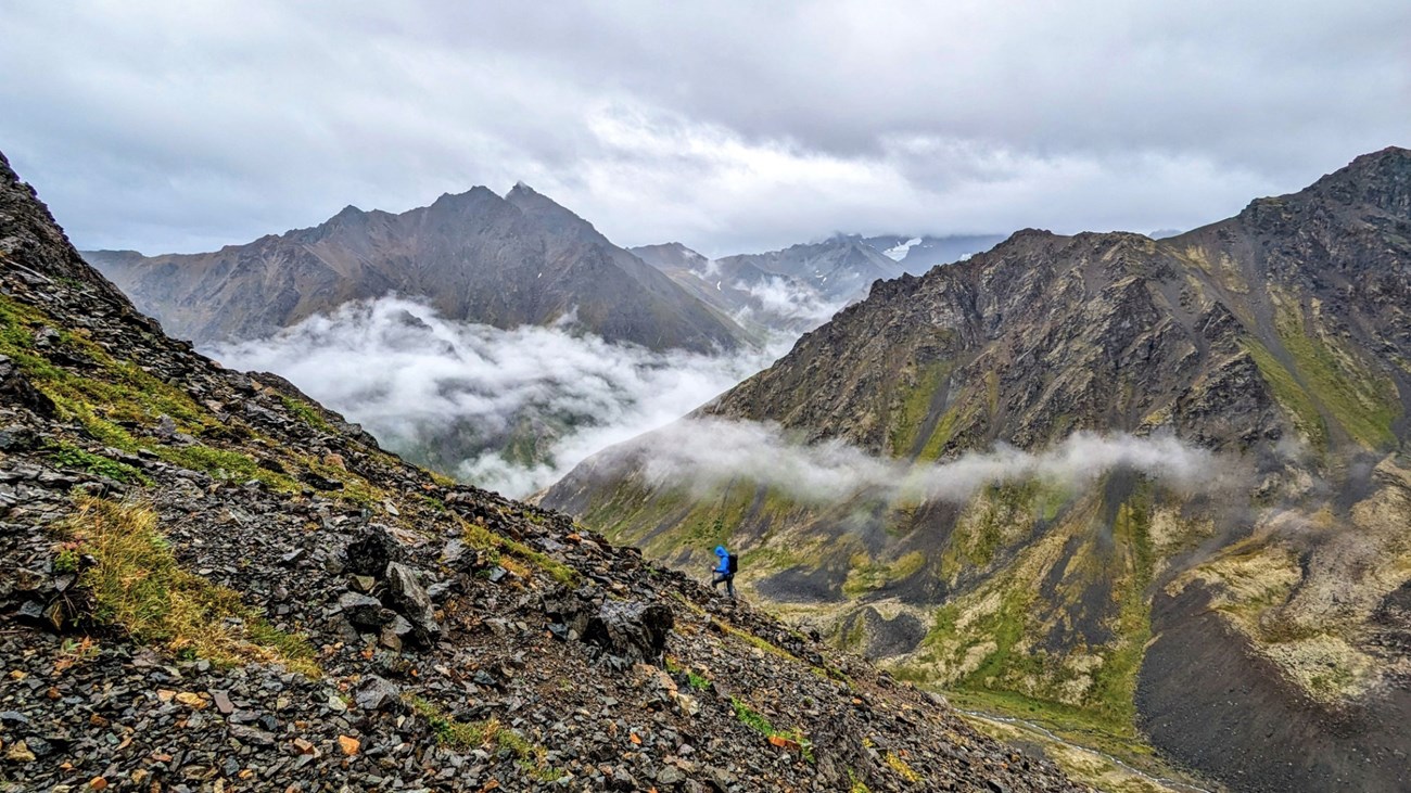 Through the Clouds A hiker wearing a blue sweatshirt hikes up a rocky mountain with patchy vegetation. They are surrounded by jagged mountains and low-hanging clouds.