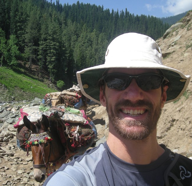 Eric Dinger Man leads a line of colorfully decorated donkeys bearing equipment on a mountain trail.