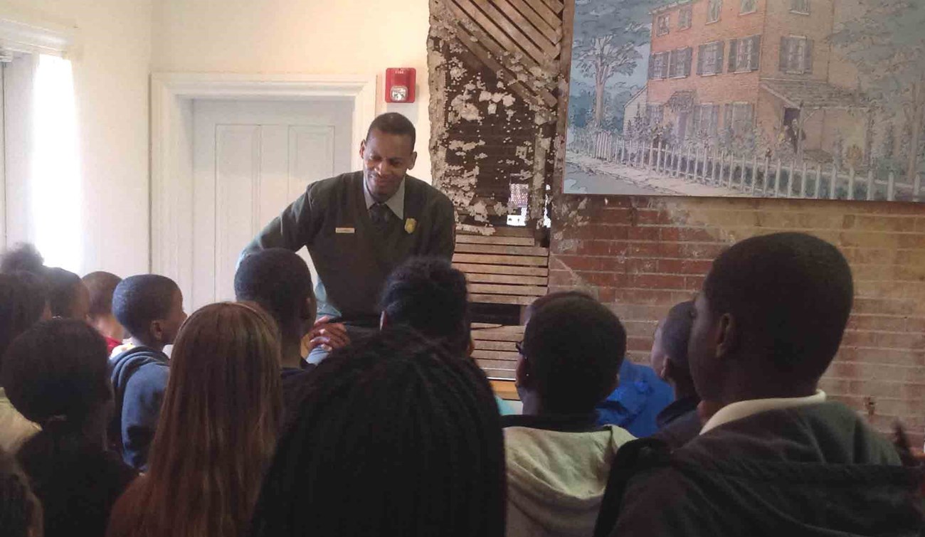 Eric Knight EDAL Park ranger talking to a large group, a painting of Edgar Allan Poe's home is hanging behind him
