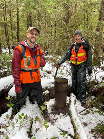 Two men in orange vests standing near an orphaned well sticking out of the ground