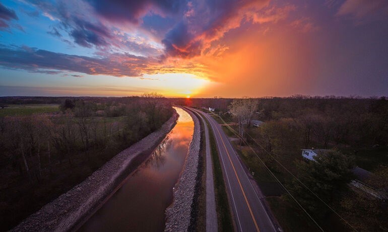 Aerial photo facing virbant sunset over horizon. Pastel and clouded sky, with sunlight reflecting on canal water in foreground. Canal recedes in distance, bordered by street on right and green field on left. Trees on both sides of canal.
