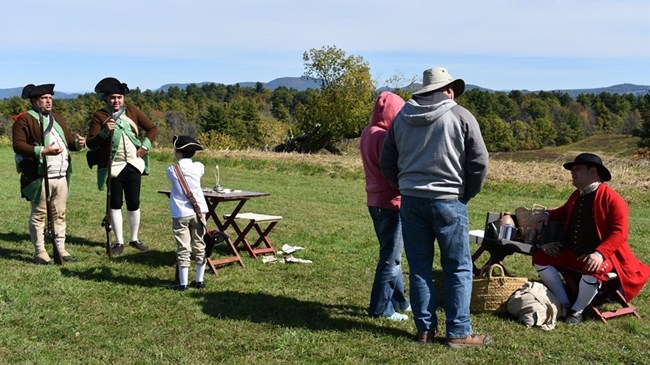 Saratoga National Historical Park At right, man and child stand in green field listening to seated man in red Revolutionary-era reproduction uniform. At left, man holding rifle in brown Revolutionary-era coat and tri-cornered hat talking with boy also in Colonial costume.
