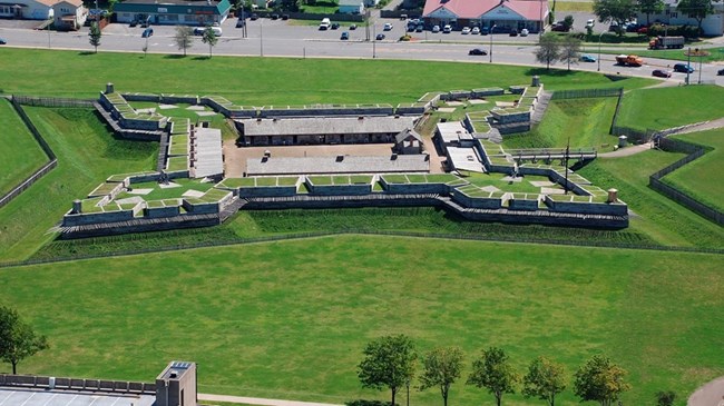 Aerial photo of elaborate fort designed with pointed diamond-shaped walls at each of the four corners of its large stone walls. All around fort are groomed bright green lawns.