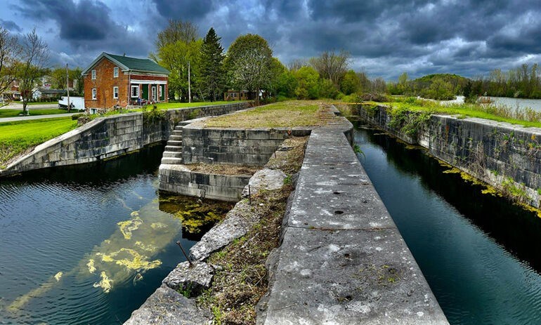 Stones canal lock area over canal water reflecting light of sky. Vibrant green grass and trees in background, red brick house with white trim in rear left, and dramatic clouds in sky.