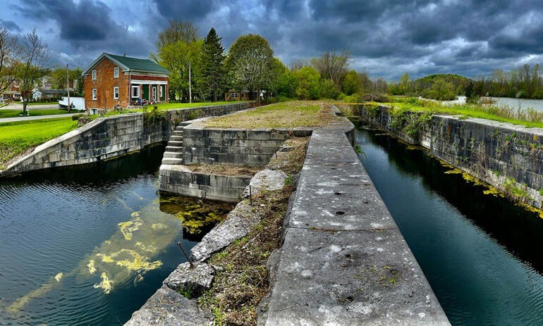 Cory Reynolds/ Erie Canalway NHC Stones canal lock area over canal water reflecting light of sky. Vibrant green grass and trees in background, red brick house with white trim in rear left, and dramatic clouds in sky.