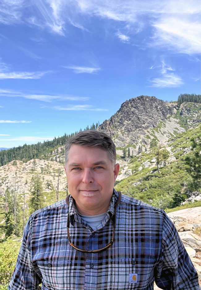 A person with short, cropped dark and gray hair, smiles as they sit with their back to a vast mountain view.