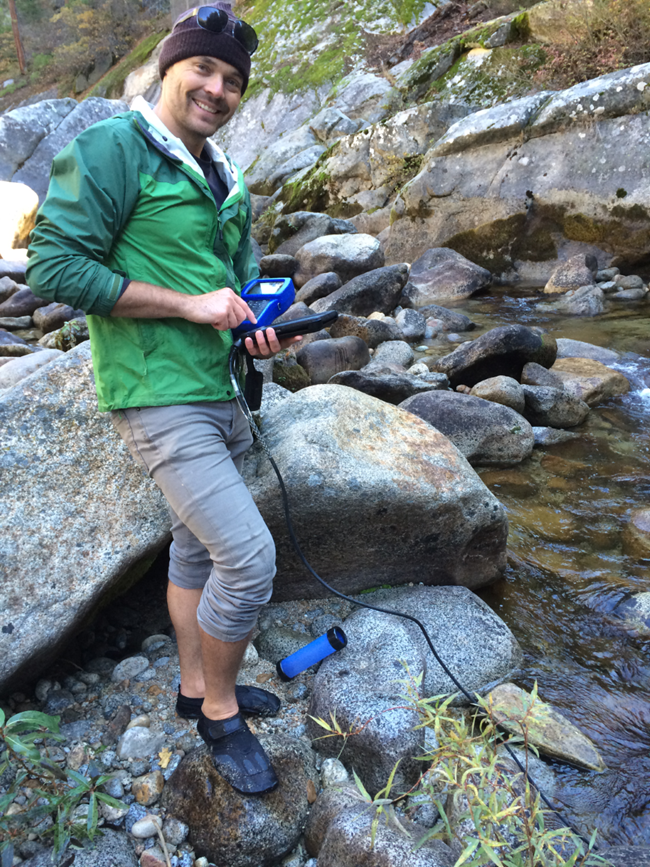 Man stands at the edge of a creek, holding an instrument that takes water quality measurements.