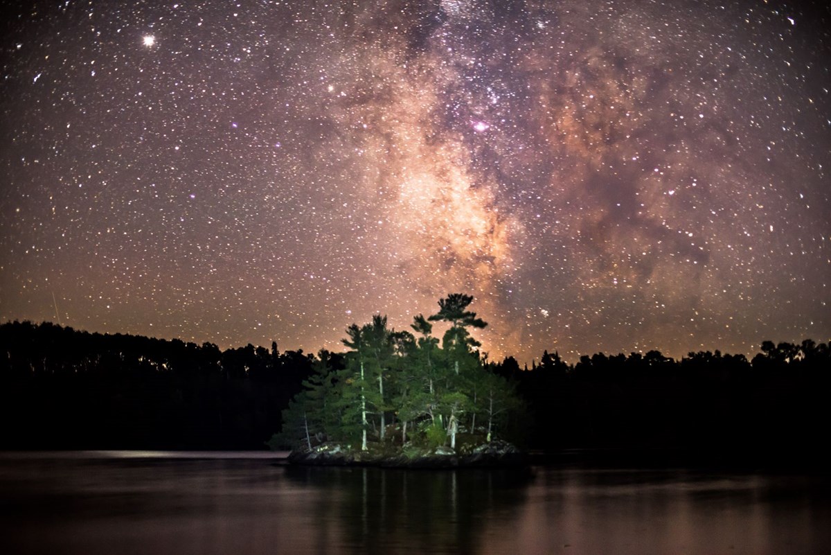 A small island covered in evergreen trees is illuminated by a sky full of stars.  The milky way streaks down the center of the night sky.  Both the sky and lake have a purple and pink hue.
