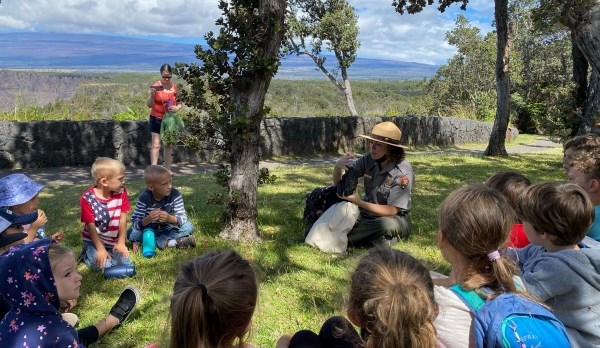 A group of young children sit in a circle with a park ranger at a grassy overlook.