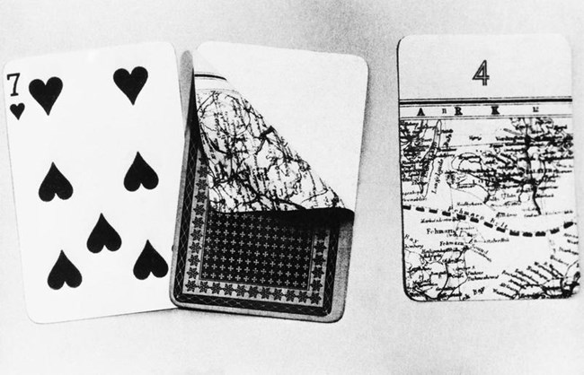 Black & white photo of 3 playing cards. The left card is a 7 of hearts. The back of the center card is facing up with the top layer peeled back to reveal the map underneath. The right card features part of a map with the number 4 printed at the top.