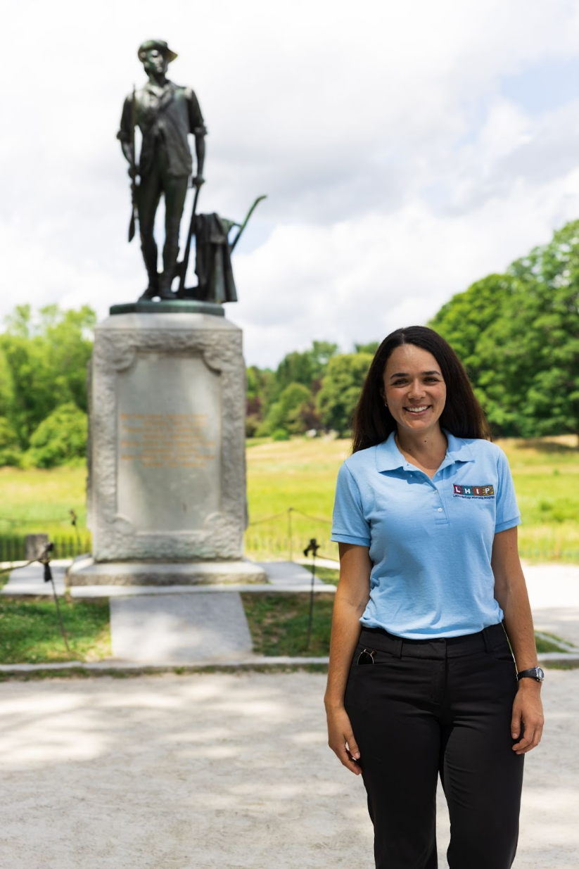 A young latina woman smiling, wearing a blue polo shirt, with colorful letters that spell "LHIP." She is wearing black pants. Behind her is a black statue of a military person, green trees, grass, and cloudy sky.