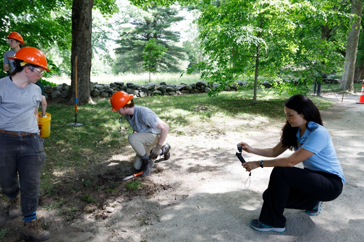 A young latina woman in a light blue polo shirt, dark pants, and sneakers holding up a measuring tool, while three other crew members in bright orange hard hats, gray shirts, and dark or khaki pants work on a trail.