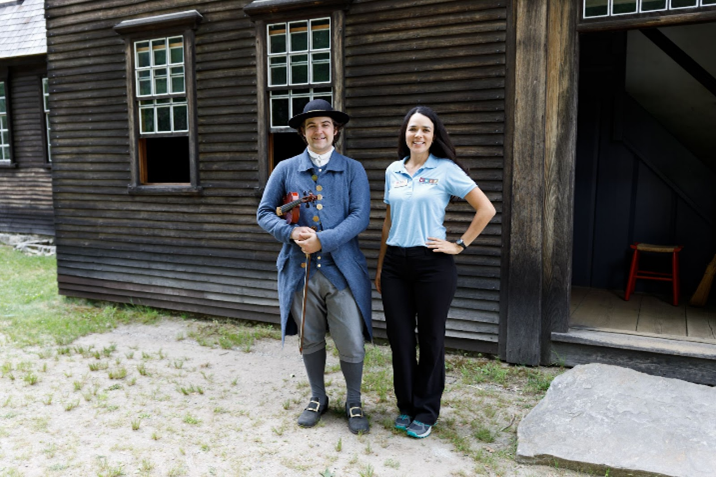 A young latina woman wearing a light blue polo shirt with the letters "LHIP" in different colors, dark pants, and sneakers, poses with her left hand on hip. She is standing next to a male dressed as an eighteenth century New England farmer