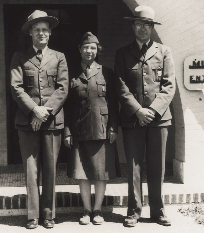 Three uniformed rangers stand in front of a stucco building.  The middle person is a woman in a skirt and flight cap while the men on either side of her wear stetson style hats. They stand with their hands clasped.
